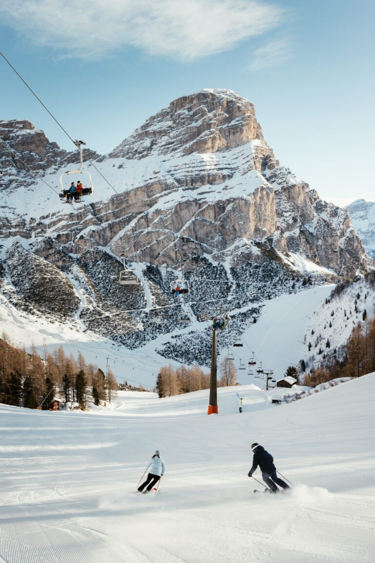 Two skiers carving down a groomed piste in Alta Badia beneath a chairlift and a towering Dolomite peak