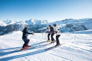 Maison Sport instructor leading beginner ski lessons for a group on a sunny piste with Alpine peaks behind