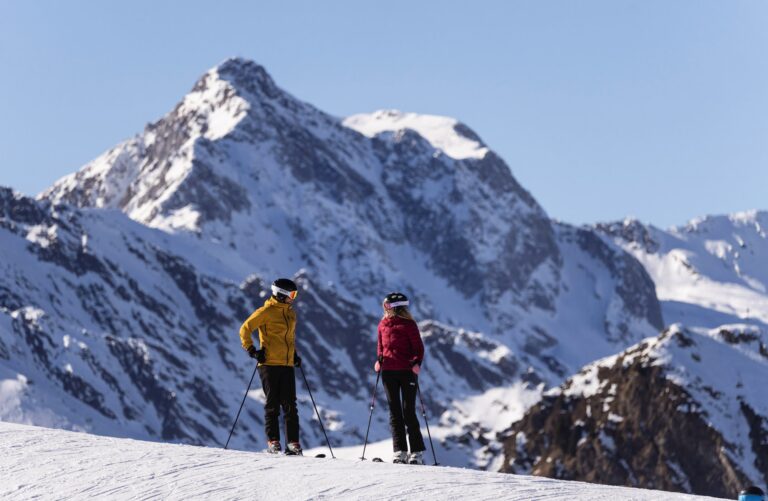 Two skiers chatting on a sunny piste in Ratschings-Jaufen with a snowy mountain peak behind