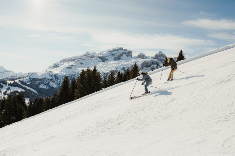 Couple skiing a steep groomed slope in Alta Badia with snowy Dolomite peaks behind