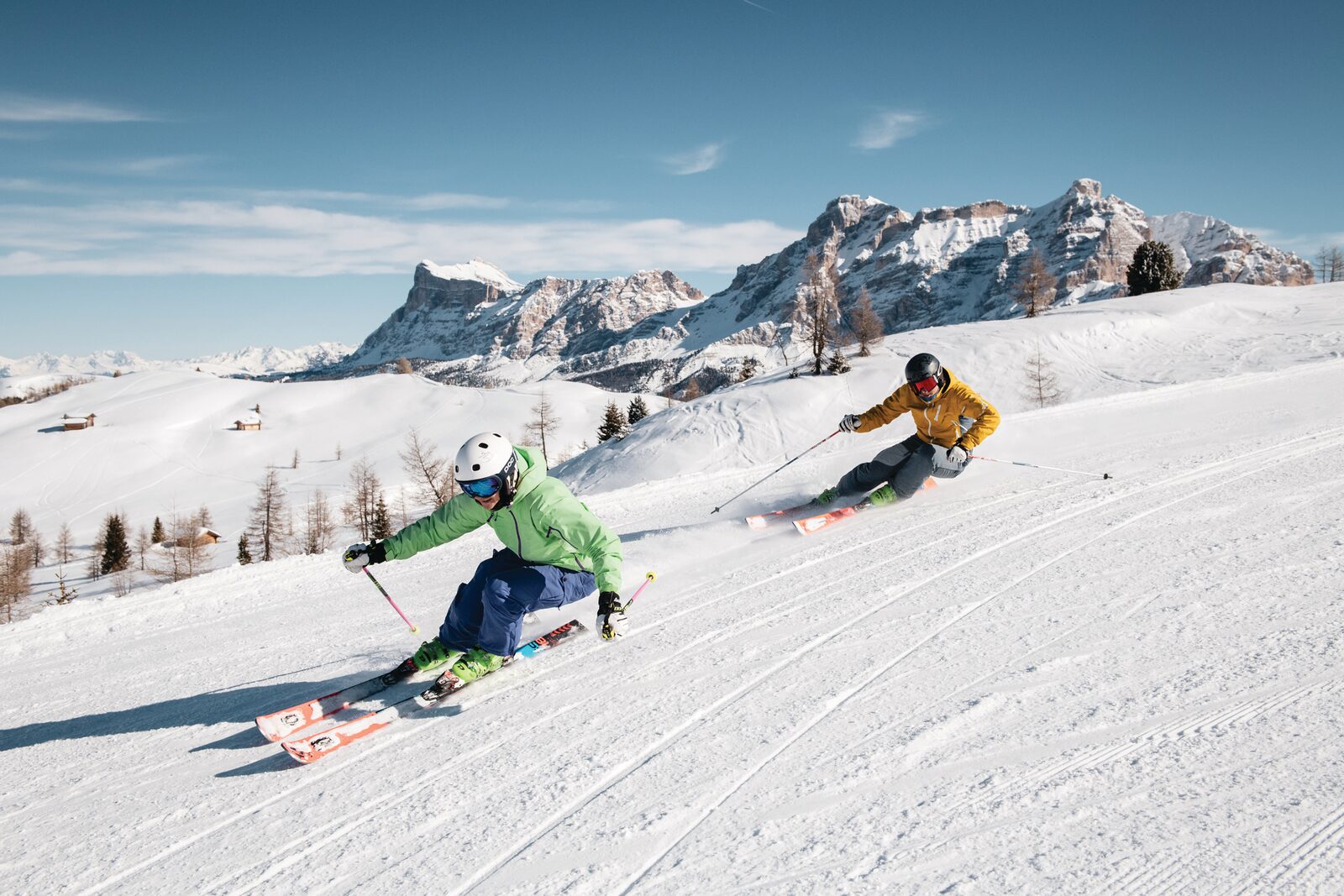 Two skiers carving an empty groomed piste in Alta Badia with Dolomites peaks behind
