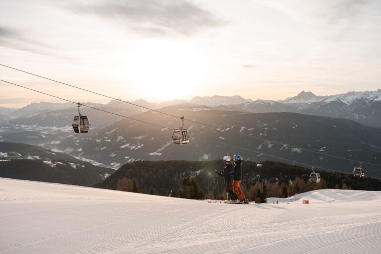 Two skiers watching the sun set over the Dolomites beneath the Gitschberg Jochtal gondola