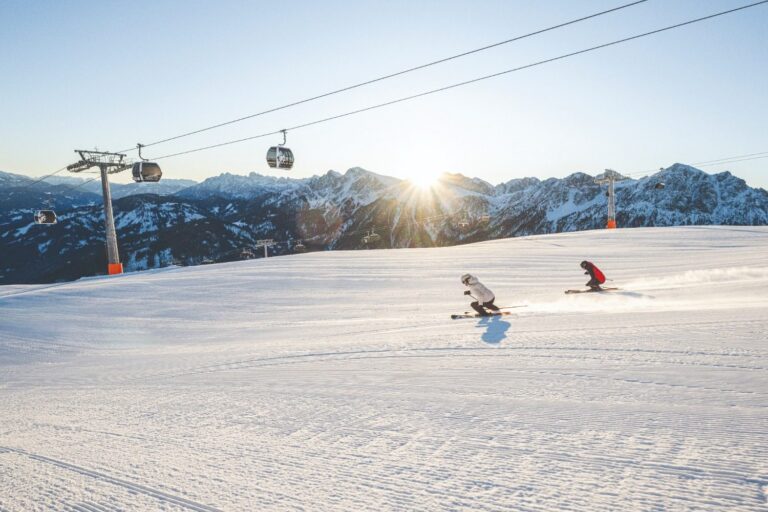 Two skiers carving across a freshly groomed piste beneath the gondola on Kronplatz at sunrise