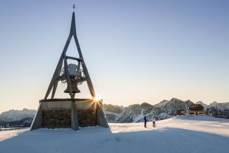 The Concordia peace bell on the Kronplatz summit at sunrise with skiers nearby
