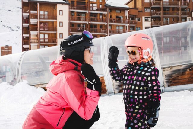 Mother and daughter high-five on the snow beside slope-side apartments in Les Menuires