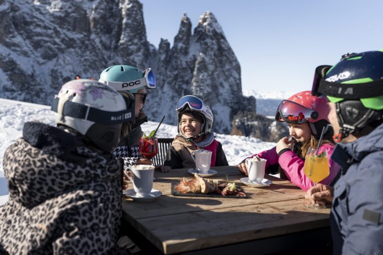 Family enjoying hot chocolate and a South Tyrolean platter on a sunny hut terrace on the Seiser Alm