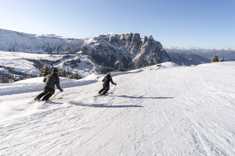 Two skiers carving down a wide piste on the Seiser Alm towards the Schlern