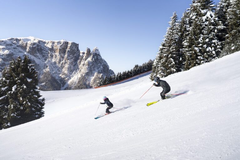 Two skiers on a tree-lined piste on the Seiser Alm with the Schlern massif behind